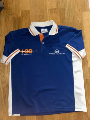 Blå piké från Sergio Tacchini - Säljer en blå pikétröja från Sergio Tacchini med vita och orange detaljer. Tröjan har vit krage, vita sidor och ärmslut med orange ränder. Tryck på bröstet och knappslå framtill. Perfekt för dig som gillar sportig stil.