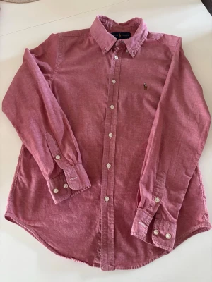 Rosa linneskjorta från Ralph Lauren - Säljer en klassisk rosa skjorta från Ralph Lauren i linne. Skjortan har button-down krage, vita knappar framtill och på manschetterna samt den ikoniska broderade loggan på bröstet. Perfekt för dig som gillar stilrena och tidlösa plagg.(L I BARNSTORLEK SÅ EJ L I HERR) Skriv privat för mer bilder.