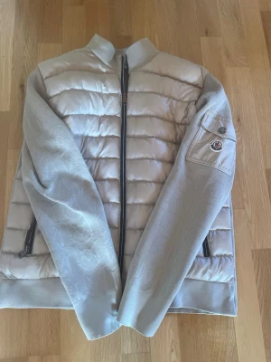 Beige pufferjacka från Moncler - Säljer en beige pufferjacka från Moncler med quiltad front och släta ärmar. Jackan har dragkedja framtill, två sidofickor med dragkedja och en ficka med Moncler-logga på ärmen. Materialet är en mix av vadderad nylon och strukturerad textil på ärmar och rygg.