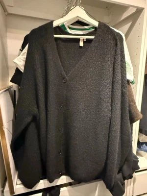 Mörkgrå kofta från Nelly - Mysig mörkgrå kofta från Nelly med v-ringning och knäppning framtill. Koftan har en lös passform och långa ärmar, perfekt att slänga över en t-shirt eller topp. Materialet är mjukt och fluffigt för extra komfort. Helt ny