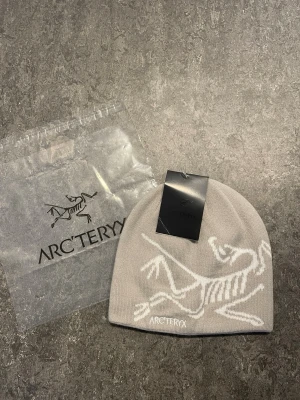 Ljusgrå mössa från Arc'teryx - Snygg ljusgrå mössa från Arc'teryx med vitt skelettmönster och broderad logga framtill. Tillverkad i mjukt stickat material som sitter skönt på huvudet. Perfekt för dig som gillar stilrena accessoarer med outdoor-vibe.