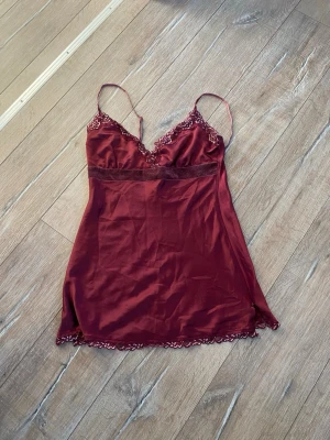 Vinrött linne med spetsdetaljer  - Snyggt vinrött linne i satin med tunna axelband och spetsdetaljer vid urringning och nederkant. Modell med slits på sidan, perfekt för en lyxig känsla. Materialet känns mjukt och glansigt mot huden. Linnet är från esprit.