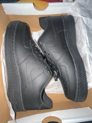 Nike Air Force 1 helsvarta sneakers - Säljer ett par klassiska Nike Air Force 1 sneakers i helsvart läder. Skorna har den ikoniska swoosh-loggan på sidan, perforerad tåbox och tjock sula med mönstrad undersida. Perfekta för dig som gillar stilrena och tidlösa sneakers.