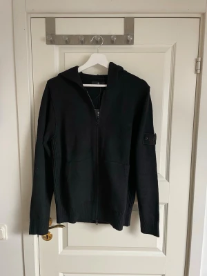 Svart hoodie från Stone Island - Säljer en svart hoodie från Stone Island med dragkedja framtill och två fickor. Hoodien har ribbade muddar och en klassisk patch på ärmen. Materialet är stickad bomull som känns både mjuk och exklusiv. Perfekt för dig som gillar streetwear och snygga detaljer.