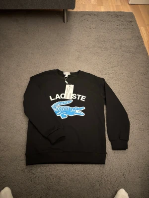 Svart sweatshirt från Lacoste - Svart sweatshirt från Lacoste med stor blå krokodil och vit logga på bröstet. Tröjan har rund halsringning, ribbade muddar och är i regular fit. Perfekt för en stilren och sportig look.