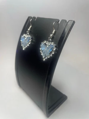 Blå hjärtformade örhängen med fjäril - Säljer ett par unika örhängen med hjärtformad design i blått och silver. I mitten av varje hjärta finns en detaljerad blå fjäril och runt kanten sitter små pärlliknande detaljer. Perfekta för dig som gillar söta och iögonfallande accessoarer.