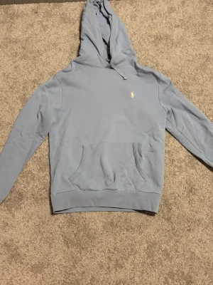 Ljusblå hoodie från Polo Ralph Lauren - Snygg ljusblå hoodie från Polo Ralph Lauren med klassisk huva och känguruficka framtill. Liten broderad logga i vitt på bröstet ger en clean look. Tillverkad i mjukt bomullsmaterial som är skönt att ha på sig. Perfekt för en avslappnad och stilren outfit.
