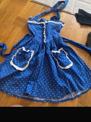 Blå prickig klänning från Hell Bunny - Supersöt blå kortklänning från Hell Bunny med vita prickar, volangkant och dekorativa rosetter på fickorna. Klänningen har knytband i midjan och axelband samt spetsdetaljer runt fickorna. Perfekt för dig som gillar retrostil!