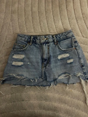 Ljusblå jeanskjol från Bershka - Trendig ljusblå jeanskjol från Bershka med slitna detaljer och fransig nederkant. Klassisk femficksmodell med bälteshällor och knappgylf. Perfekt för en avslappnad och cool stil. Ganska liten i storleken!