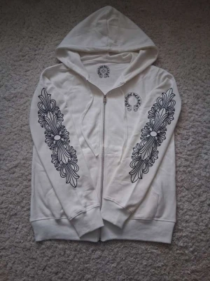 Vit hoodie från Chrome Hearts - Säljer en vit hoodie från Chrome Hearts med dragkedja och stora svarta grafiska tryck längs båda ärmarna samt logga på bröstet och ryggen. Hoodien har huva och ribbade muddar. Perfekt för dig som gillar streetwear och unika detaljer.