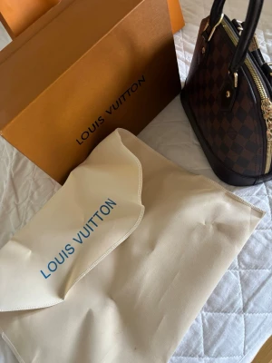 Louis Vuitton Damier Alma BB handväska - Säljer en Louis Vuitton Alma BB handväska i klassiskt brunt Damier-mönster. Väskan har rundad form, dubbla handtag i mörkbrunt läder och guldfärgade metalldetaljer. Kommer med dustbag och originalkartong. Perfekt för dig som gillar lyxiga accessoarer. Säljer då den inte kommer till användning längre! PRIS KAN DISKUTERAS!!!
