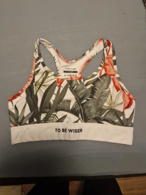Sport-bh med tropiskt mönster To Be Wiser - Snygg sport-bh från To Be Wiser med tropiskt bladmönster i grönt, vitt och orange. Bh:n har brottarrygg och bred resår med texten 'TO BE WISER' framtill. Perfekt för träning och aktiviteter där du vill ha både stil och stöd.