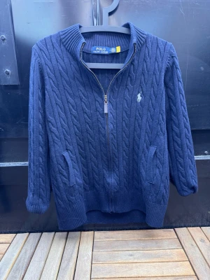 Mörkblå kabelstickad tröja Polo Ralph Lauren🙌🤩 - Mörkblå kabelstickad tröja från Polo Ralph Lauren med dragkedja framtill, hög krage och två fickor. Klassisk vit logga broderad på bröstet. Tröjan har ribbade muddar och är perfekt för dig som gillar en stilren look.🙌🤩