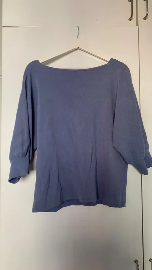 Blå topp från KappAhl - Säljer en blå topp från KappAhl med lös passform och trekvartsärmar. Toppen har en bred, båtringad hals och är tillverkad i ett mjukt, ribbat material som känns skönt mot huden. Perfekt för en avslappnad och stilren look.