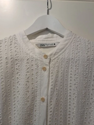 Vit broderad klänning från Zara med skärp - Vit klänning från Zara i broderad bomull med knappar framtill och knytskärp i midjan. Klänningen har långa ärmar och ett vackert hålmönster som ger en luftig och elegant känsla. Klänningen är fodrad med tunnt vitt bomullstyg Perfekt för varma dagar och stilrena outfits. Endadt använd på studentdagen och därefter tvättad.