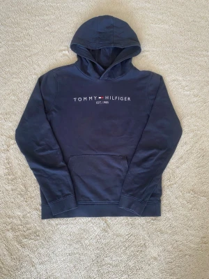 Marinblå hoodie Tommy Hilfiger - Snygg marinblå hoodie från Tommy Hilfiger med klassisk logga och text 'EST. 1985' på bröstet. Hoodien har stor huva, magficka och ribbade muddar. Tillverkad i mjuk bomullsblandning som känns skön mot huden. Perfekt för en avslappnad och stilren look.