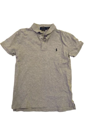 Grå pikétröja från Polo Ralph Lauren - Klassisk grå pikétröja från Polo Ralph Lauren med korta ärmar och krage. Tröjan har två knappar vid halsen och den ikoniska broderade loggan på bröstet. Tillverkad i mjuk bomull för en bekväm känsla. Passar runt 160-167cm lång, pris kan diskuteras, hör av er vid fler frågor eller funderingar.