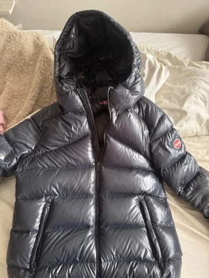 Mörkblå dunjacka från Colmar - Snygg mörkblå dunjacka från Colmar med glansig finish och stor huva. Jackan har två dragkedjeförsedda fickor framtill och Colmar-logga på ärmen. Perfekt för kalla vinterdagar och riktigt trendig look.