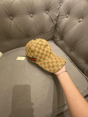 Gucci keps med GG-mönster - Snygg beige keps från Gucci med klassiskt GG-monogram i brunt över hela kepsen. På sidan finns den ikoniska gröna och röda randen. Kepsen har böjd skärm och justerbar rem bak. Perfekt för dig som vill ha en lyxig streetstyle-look. Du får boxen med.