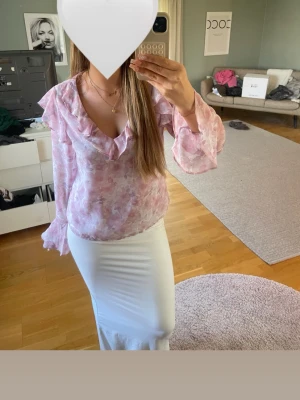 Ljusrosa blommig volangblus - Blus i ljusrosa med blommigt mönster och volangdetaljer längs v-ringningen. Långa, transparenta ärmar och luftig passform. 