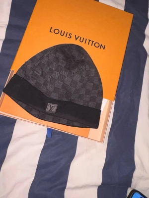 Autentisk Louis Vuitton Mössa - Snygg svart mössa från Louis Vuitton med diskret rutmönster och klassisk LV-logga framtill. Mössan har en bredare kant nertill och är tillverkad i ett mjukt material som sitter skönt på huvudet. Perfekt accessoar för att lyfta din streetwear-look. (Köpt från Louis Vuittons hemsida)