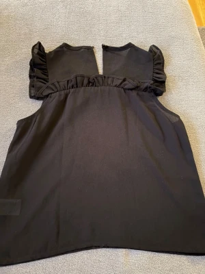 Svart ärmlös topp med volang  - Snygg svart ärmlös topp  med volangdetalj över axlarna och runt halsen. Toppen har en rak passform och rund halsringning. Perfekt att styla med jeans eller kjol för en trendig look.