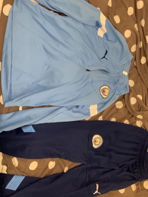 Manchester City träningsset från Puma med keps - Säljer ett Manchester City träningsset från Puma med ljusblå långärmad tröja och mörkblå byxor. Tröjan har halv dragkedja, klubbmärke och Puma-logga. Byxorna har resår i midjan, klubbmärke och detaljer i ljusblått. Perfekt för fotbollsträning. Aldrig använt bara tvättat