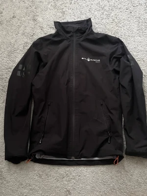 Svart Gore-Tex regnjacka Sail Racing - Svart regnjacka från Sail Racing i Gore-Tex-material. Jackan har hel dragkedja framtill, två sidofickor med dragkedja och diskreta loggor på bröstet och ärmen. Perfekt för blöta dagar och snygg minimalistisk design.