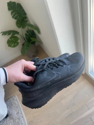 Svarta löparskor Nike  - Säljer ett par helsvarta löparskor ifrån Nike med låg profil och tjock, strukturerad sula. Skorna har en diskret Nike-logga på sidan, snörning och är tillverkade i ett slitstarkt textilmaterial. Perfekta för dig som gillar en clean och sportig look.