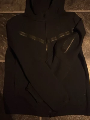 Svart Nike hoodie med dragkedja  - Säljer en svart hoodie från Nike i storlek XL. Hoodien har dragkedja framtill, huva och diskreta detaljer på bröstet. Tillverkad i mjukt material som passar perfekt för chill. Klassisk Nike-logga på etiketten.