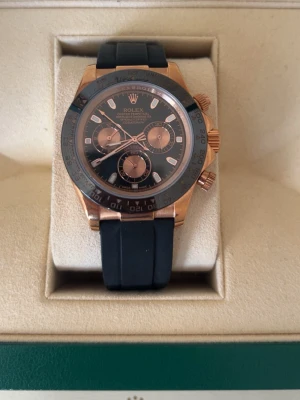 Rolx Daytona svart och guld klocka - Snygg Daytona kronograf med svart urtavla, roséguldsdetaljer och svart gummiband. Klockan har tre subdials, tydliga index och en sportig men exklusiv look. Perfekt accessoar för dig som vill sticka ut med stil.