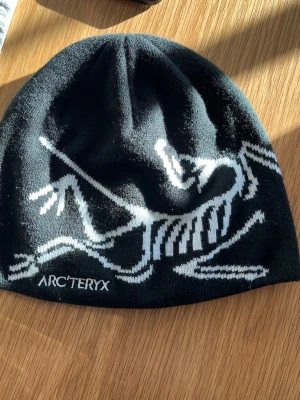 Svart mössa från Arc'teryx - ÄKTA Snygg svart mössa från Arc'teryx med vit logga och coolt skelettmönster på framsidan. Perfekt för kalla dagar och outdoor-aktiviteter. Mjuk och stretchig passform som sitter skönt på huvudet.