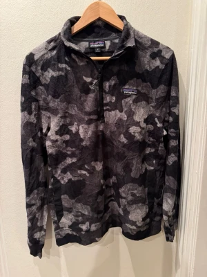 Patagonia 1/4-zip fleece camo grå/svart stl M herr - Patagonia quarter-zip i mjuk mikrofleece med botaniskt camo-mönster i grått och svart. Stl muddar och nederkant, bröstlogga. Aldrig använd, utan lapp. Helt felfri. Stl M herr.