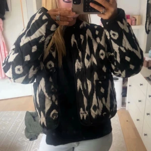 Svartvit quiltad jacka med mönster - Quiltad jacka i svart och vitt med grafiskt mönster. Jackan har knappar framtill, raka ärmar och en loose passform. Perfekt för dig som vill sticka ut med en unik och trendig look.