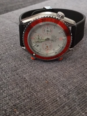 Omega Seamaster klocka med röd bezel - Snygg Omega Seamaster klocka med vit urtavla, röd bezel och svarta detaljer. Klockan har kronograf-funktion och svart armband i syntetmaterial. Silverfärgat boett och tydliga indexmarkeringar ger en sportig och stilren look.automatik