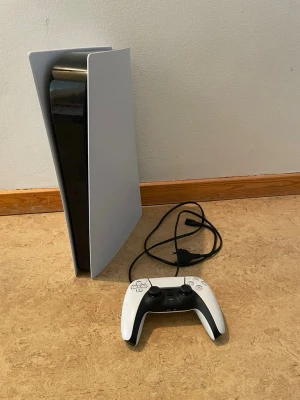 PlayStation 5 med handkontroll - Säljer en PlayStation 5 från Sony med en vit och svart DualSense-handkontroll och strömkabel. Konsolen har en futuristisk design med vita sidopaneler och svart mittdel i plast. Perfekt för gaming och nästa generations spelupplevelser.