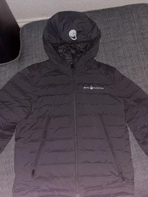 Svart dunjacka från Sail Racing - Snygg svart dunjacka från Sail Racing med huva och dragkedja framtill. Jackan har två sidofickor med dragkedja och diskret logga på bröstet. Perfekt för kalla vinterdagar och har en stilren, sportig look.