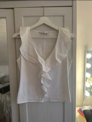 Vit ärmlös topp med volang från H&M - Säljer en vit ärmlös topp från mango med djup v-ringning och bred volang längs halsringningen och framtill. Toppen har en feminin och luftig design, perfekt för varma dagar. Tillverkad i mjukt bomullsmaterial.