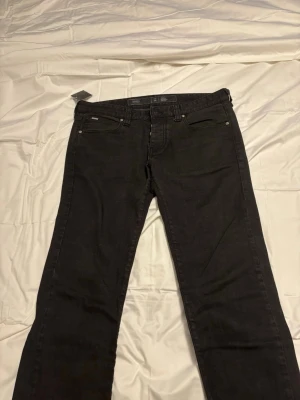 Svarta jeans från Armani - Snygga svarta jeans från Armani med klassisk femficksmodell och raka ben. Jeansen har diskreta sömmar och metallknappar, samt en liten logotyp på bakfickan. Perfekta för en stilren och enkel look. Ordinarie pris 1400kr!