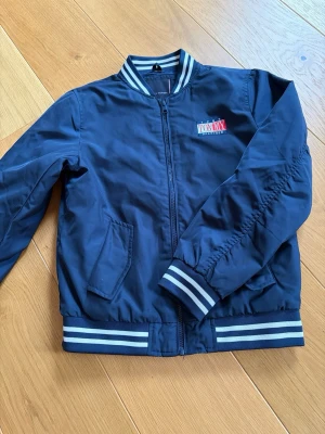 Marinblå bomberjacka Tommy Hilfiger - Snygg marinblå bomberjacka från Tommy Hilfiger med vita och blå ränder på muddar och krage. Jackan har dragkedja, två fickor fram och en broderad logga på bröstet. Tillverkad i ett slitstarkt material som passar perfekt till streetwear-stilen.