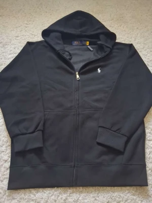 Svart hoodie från Polo Ralph Lauren - Snygg svart hoodie från Polo Ralph Lauren med dragkedja framtill och klassisk vit logga broderad på bröstet. Hoodien har huva, ribbade muddar och är tillverkad i mjukt bomullsmaterial. Perfekt för en avslappnad och stilren look.
