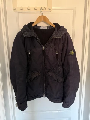 Mörkblå jacka från Stone Island - Snygg mörkblå jacka från Stone Island med huva och dragkedja framtill. Jackan har flera fickor med både dragkedja och knappar samt den klassiska logotypen på ärmen. Vid frågor eller intresse av fler bilder är det bara att höra av sig🤠