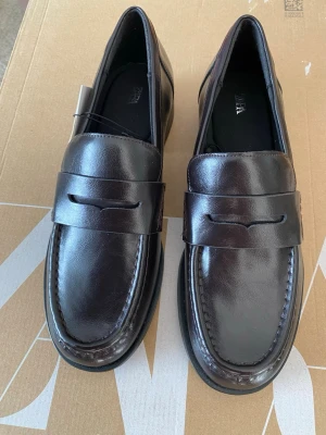 Mörkbruna loafers från Zara - Snygga mörkbruna loafers från Zara i klassisk design med slät ovandel och dekorativ rem. Skorna är tillverkade i blankt läder och har en rundad tå samt låg klack. Perfekta för dig som vill ha en stilren och tidlös look.
