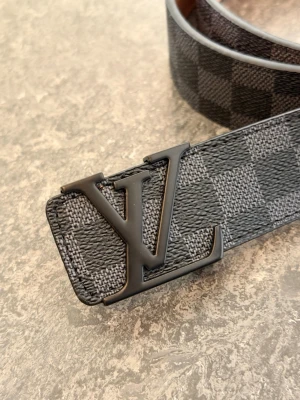 Svart rutigt LV-bälte i skinn - Snyggt svart bälte från Louis Vuitton med klassiskt rutigt mönster och stor LV-logga i metall på spännet. Bältet är gjort i skinn och har en stilren design som passar till många olika outfits.