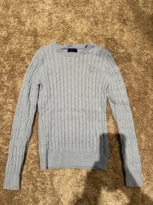 Ljusblå kabelstickad tröja Gant - Säljer en ljusblå kabelstickad tröja från Gant i stretchig bomull. Tröjan har rund halsringning, ribbade muddar och en liten broderad logga på bröstet. Perfekt för dig som gillar klassisk och stilren look.
