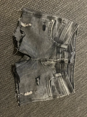 Gråa shorts  - Säljer ett par grå jeansshorts med slitna detaljer och råa kanter. Shortsen har fem fickor, knappgylf och en cool, tvättad look. Perfekta för dig som gillar en avslappnad och trendig stil.