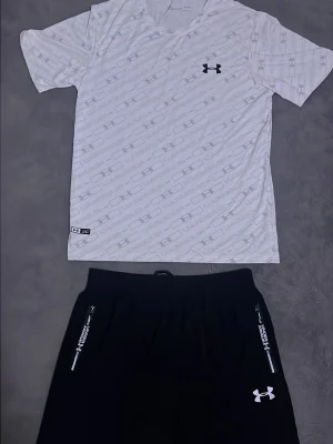 Under Armour träningsset svart/vit - Säljer ett snyggt träningsset från Under Armour med en vit t-shirt med diskret mönster och svart logga samt svarta shorts med vita detaljer och dragkedjefickor. Perfekt för gymmet eller löpning. Materialet är lätt och andas.