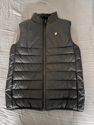 Svart quiltad dunväst Lyle & Scott - Svart dunväst från Lyle & Scott med gul logga på bröstet. Västen har quiltad design, hög krage och dragkedja framtill. Tillverkad i polyester och syntetmaterial för en lätt och bekväm känsla. Perfekt att slänga över en hoodie. Storleken: 152/158(ca: 12-13 years)