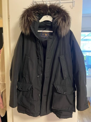 Svart parkas med äkta päls från Woolrich - Snygg svart parkas från Woolrich med stor huva och fluffig äkta päls. Jackan har flera praktiska fickor framtill och stängs med både dragkedja och knappar. Perfekt för kalla dagar och riktigt varm. Klassisk och tidlös design.