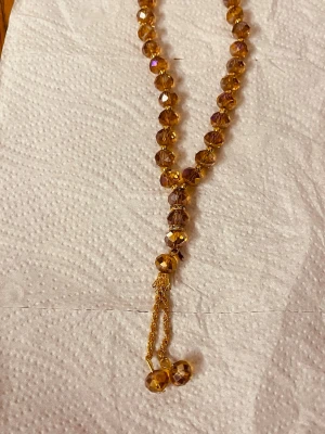 Gyllene halsband med glaspärlor - Säljer ett halsband med gnistrande gyllene glaspärlor i rund form. Halsbandet har en dekorativ tofsdetalj med två kedjor och större pärlor längst ner. Perfekt för dig som vill ha något som sticker ut och fångar ljuset snyggt.